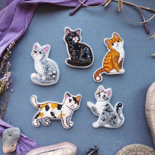 six beaded cat brooches arranged on a blue background, with some of the cats depicted in different colors and poses.