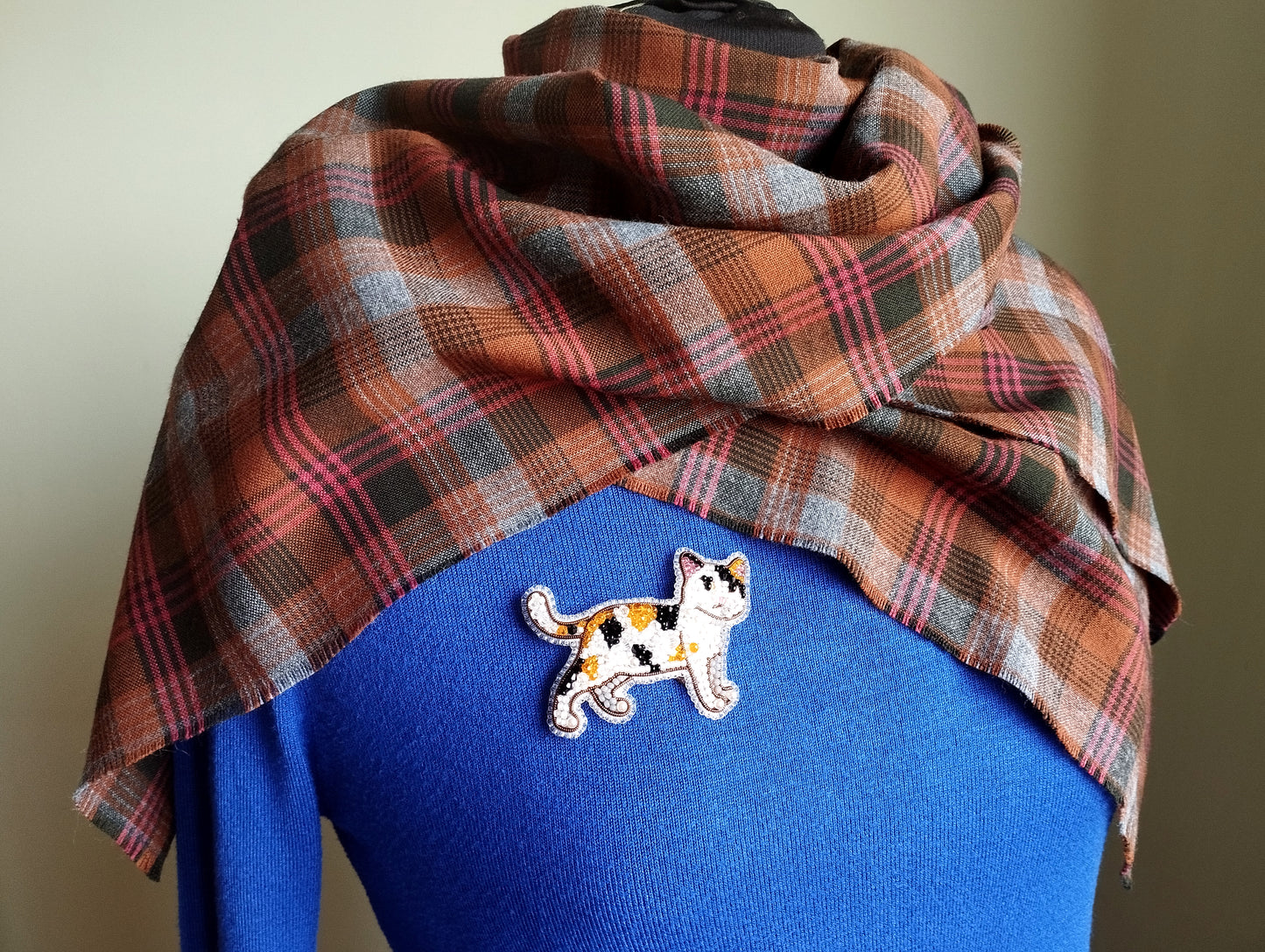 A plaid scarf is draped over a blue sweater, with a small embroidered cat pin or badge pinned to the sweater.