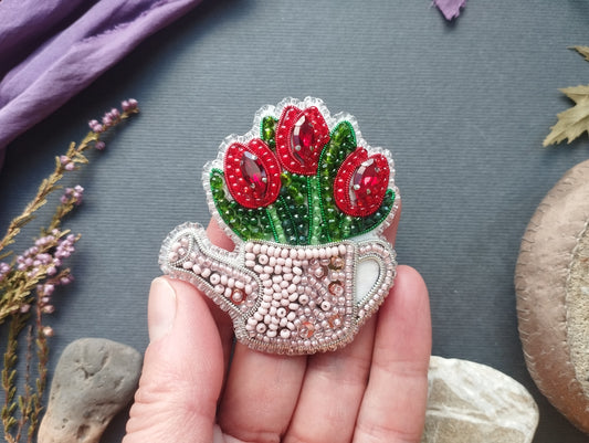 A hand holding a decorative beaded object resembling a teapot with red flowers, against a gray background with various natural elements such as leaves and rocks.