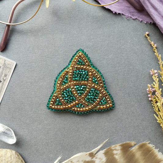 a triangular beaded object with a green and gold color scheme, placed on a gray surface surrounded by various objects such as crystals, feathers, and a card.
