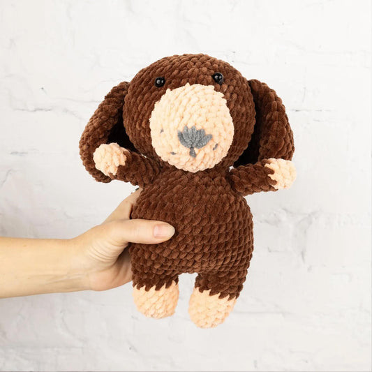 A hand is holding a brown, crocheted stuffed dog toy against a white brick wall.