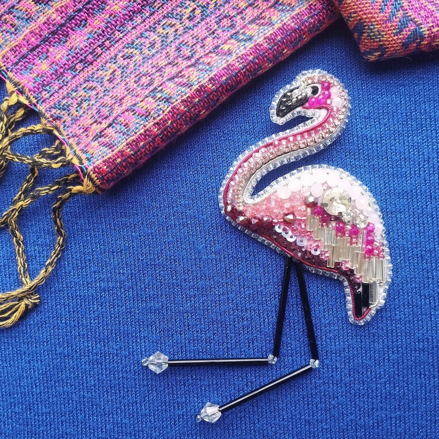 a pink flamingo embroidered on a blue fabric, with a colorful woven bag and a yellow string visible in the background.
