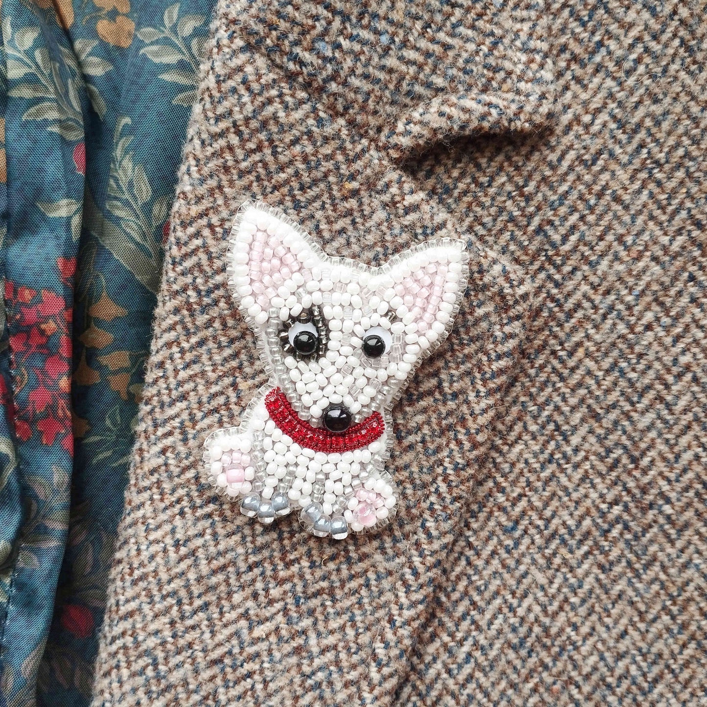 A close-up image of a small, white dog-shaped embroidered piece on a tweed fabric, with a red collar.
