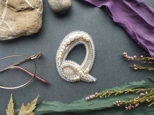 a close-up view of a decorative letter 'Q' made of beads and pearls, surrounded by various objects such as a rock, a pair of glasses, and some flowers.