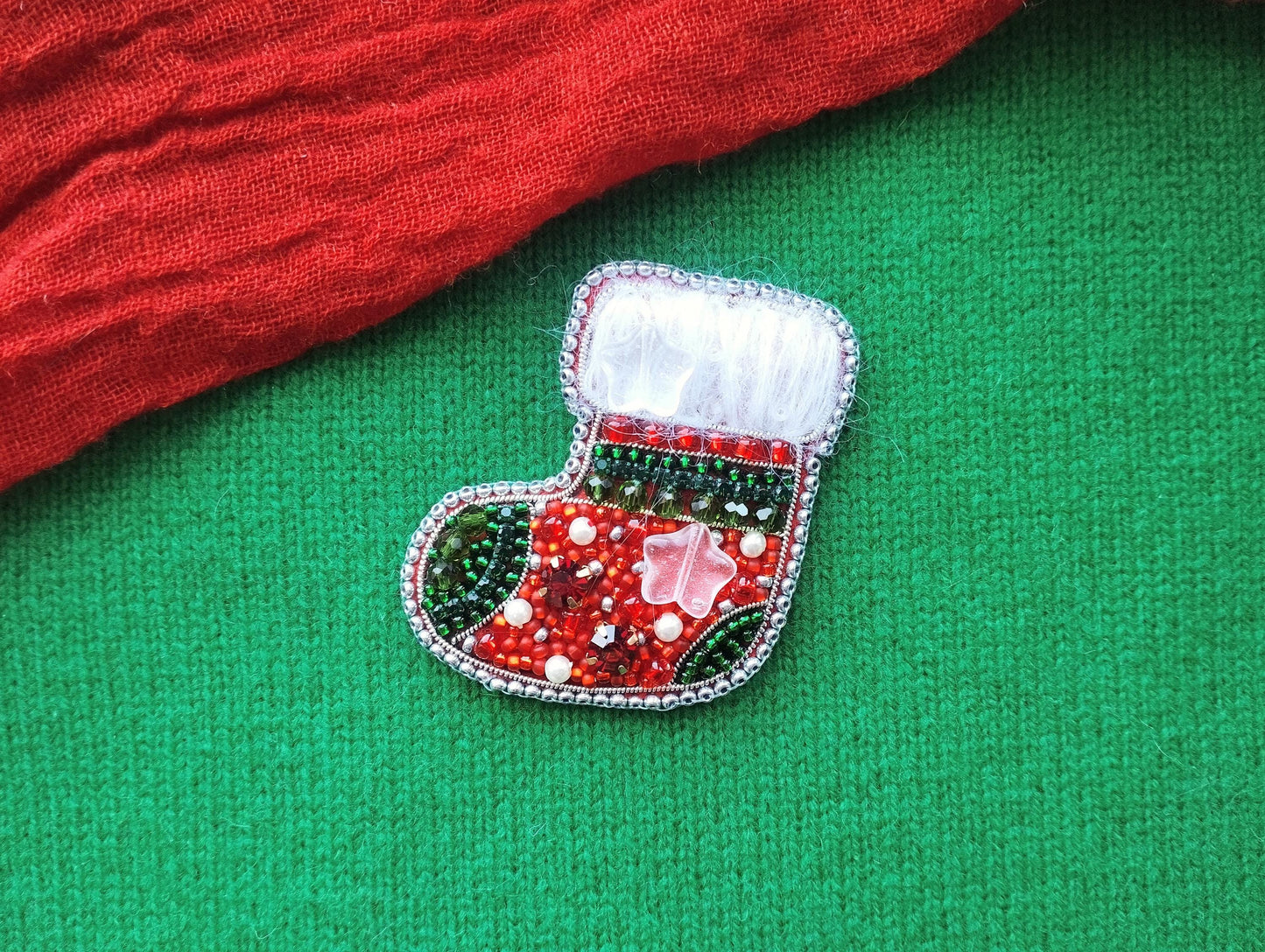 A red and green Christmas stocking ornament with beaded and embroidered details is displayed on a green fabric background.