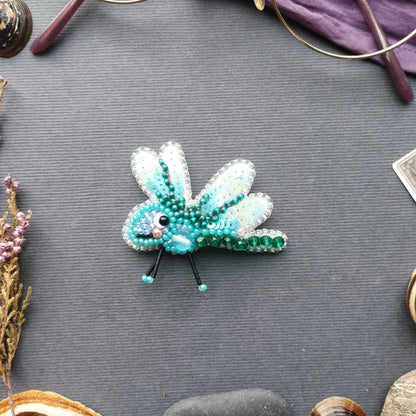 a small, intricately designed blue and green dragonfly brooch, which is placed on a grey surface surrounded by various objects.