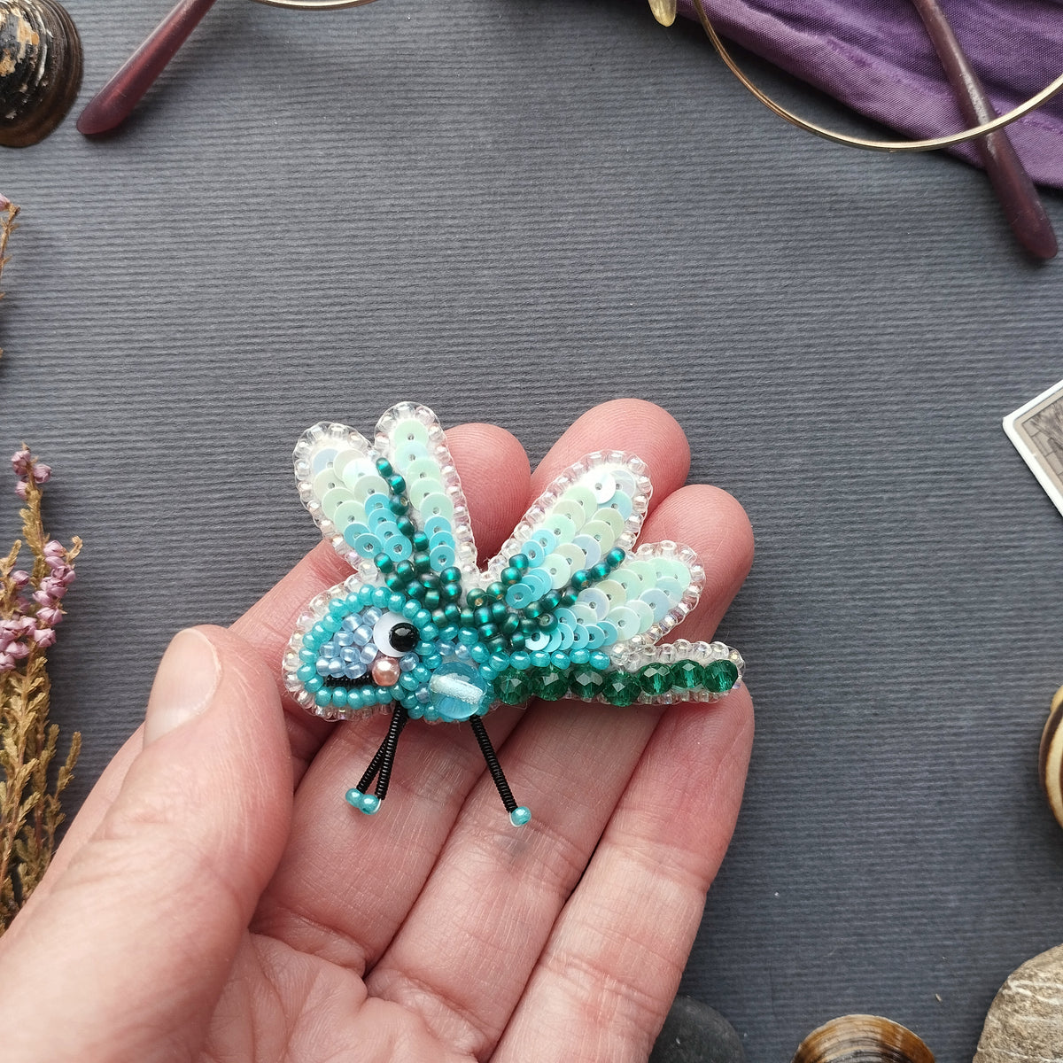 A hand holding a small, intricately beaded dragonfly brooch against a gray background.