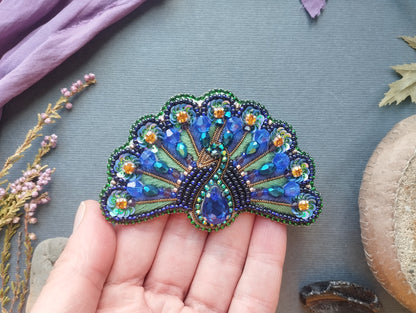 A hand holding a decorative peacock feather-shaped brooch with blue and green beads and a large blue gemstone.