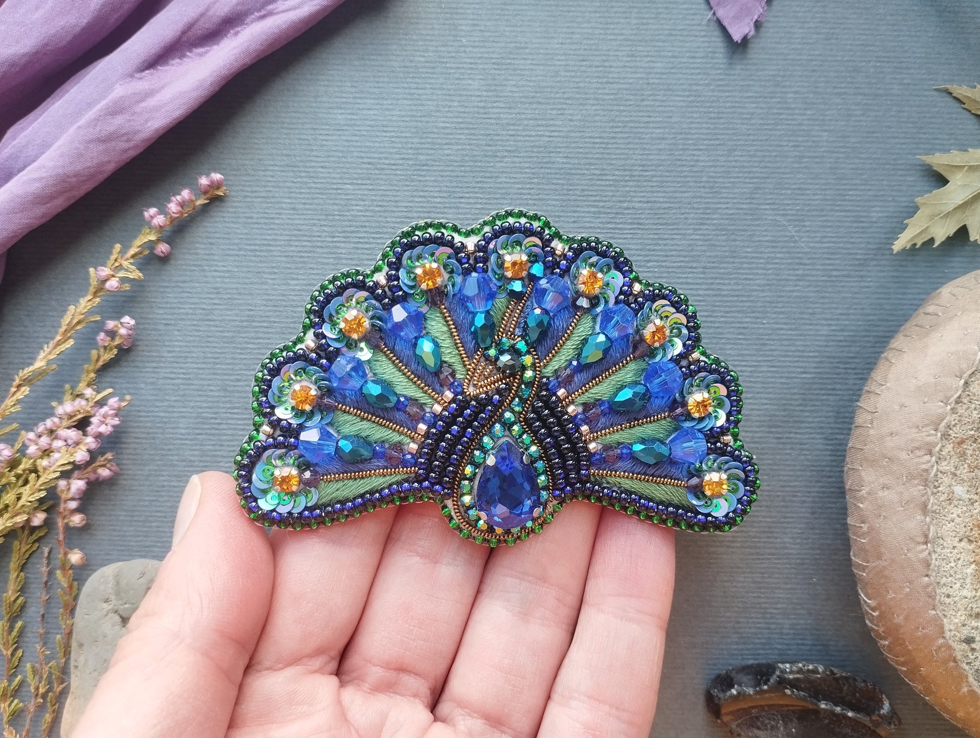 A hand holding a decorative peacock feather-shaped brooch with blue and green beads and a large blue gemstone.