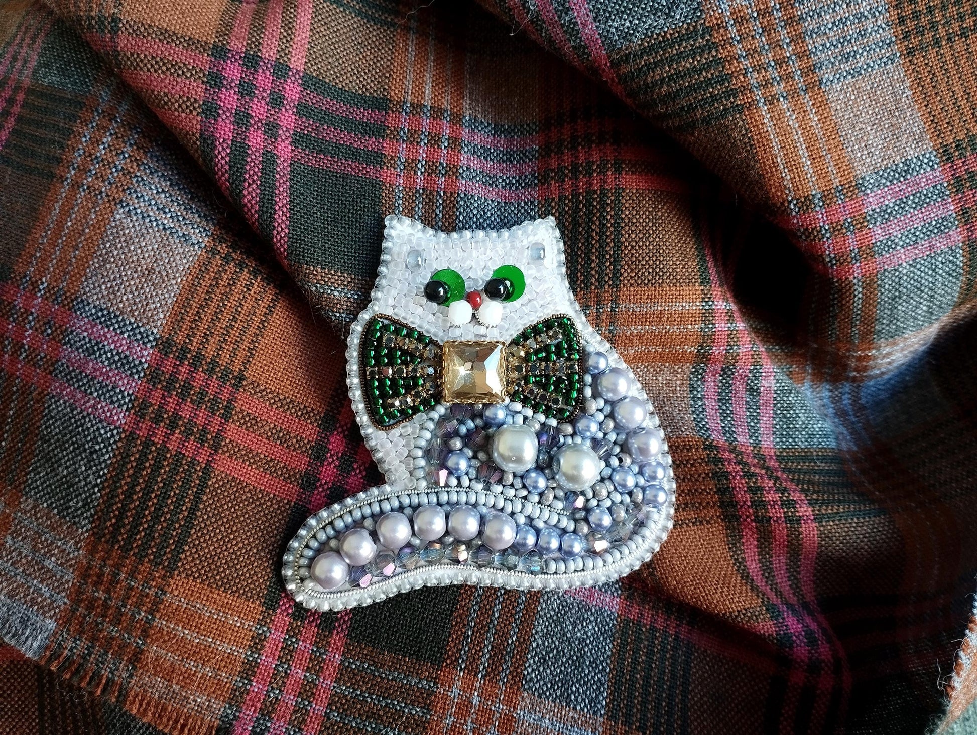 A decorative cat brooch with a green bow tie and beaded embellishments, resting on a plaid fabric background.