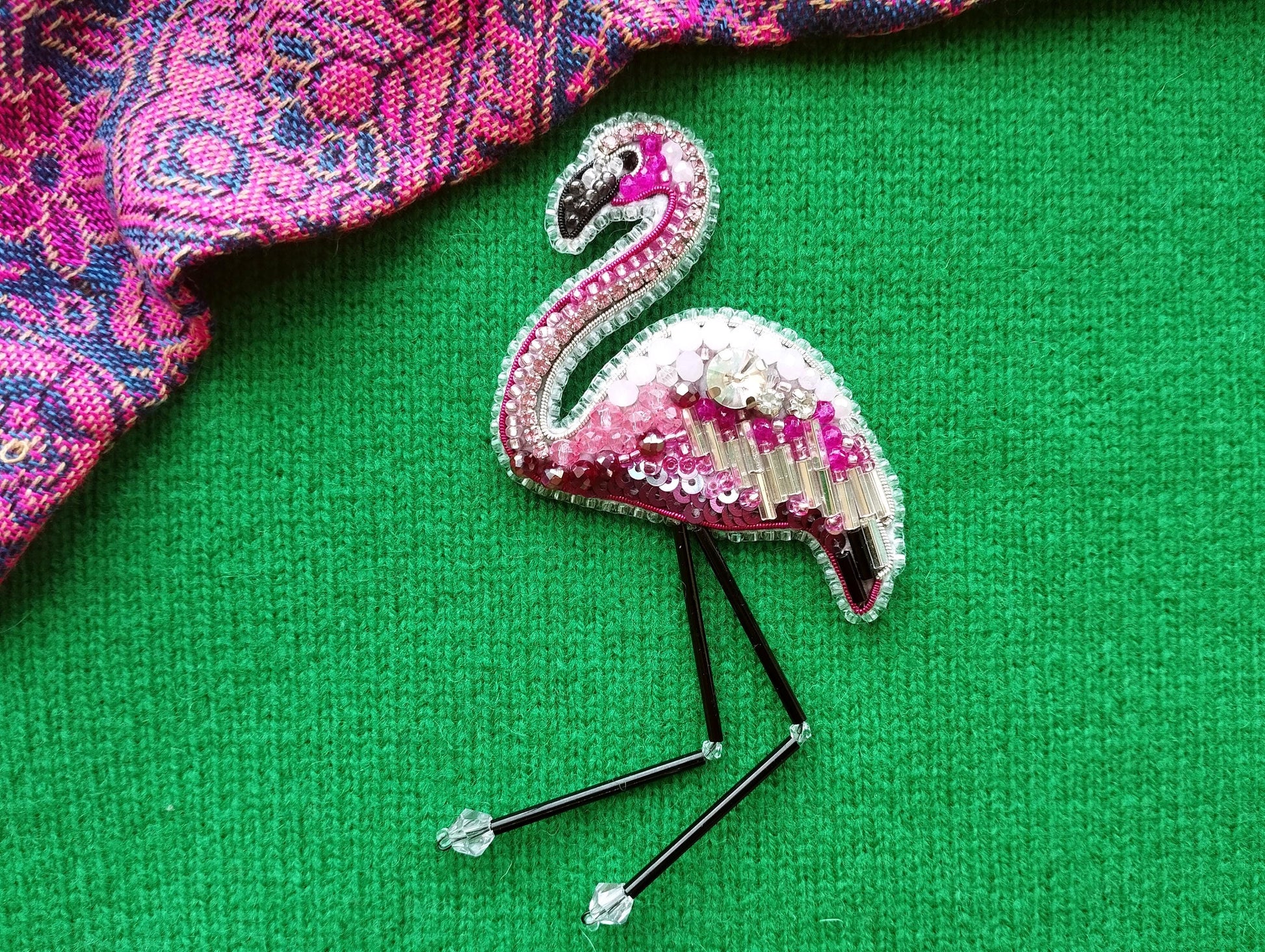 A pink and white beaded flamingo brooch is displayed on a green surface, with a patterned fabric visible in the background.