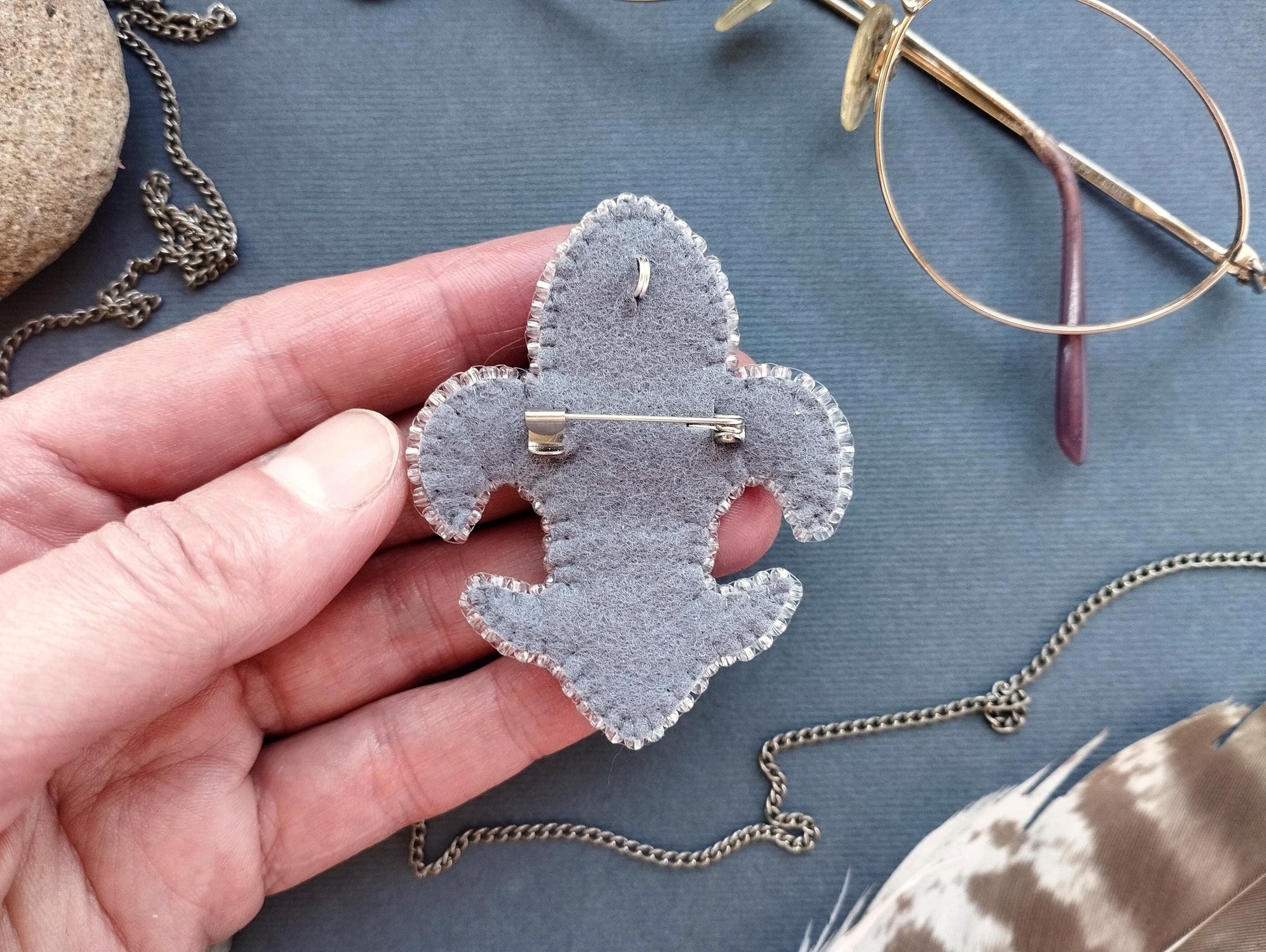 A hand holding a gray felt badge with a pin, surrounded by various accessories such as glasses, a chain, and feathers.