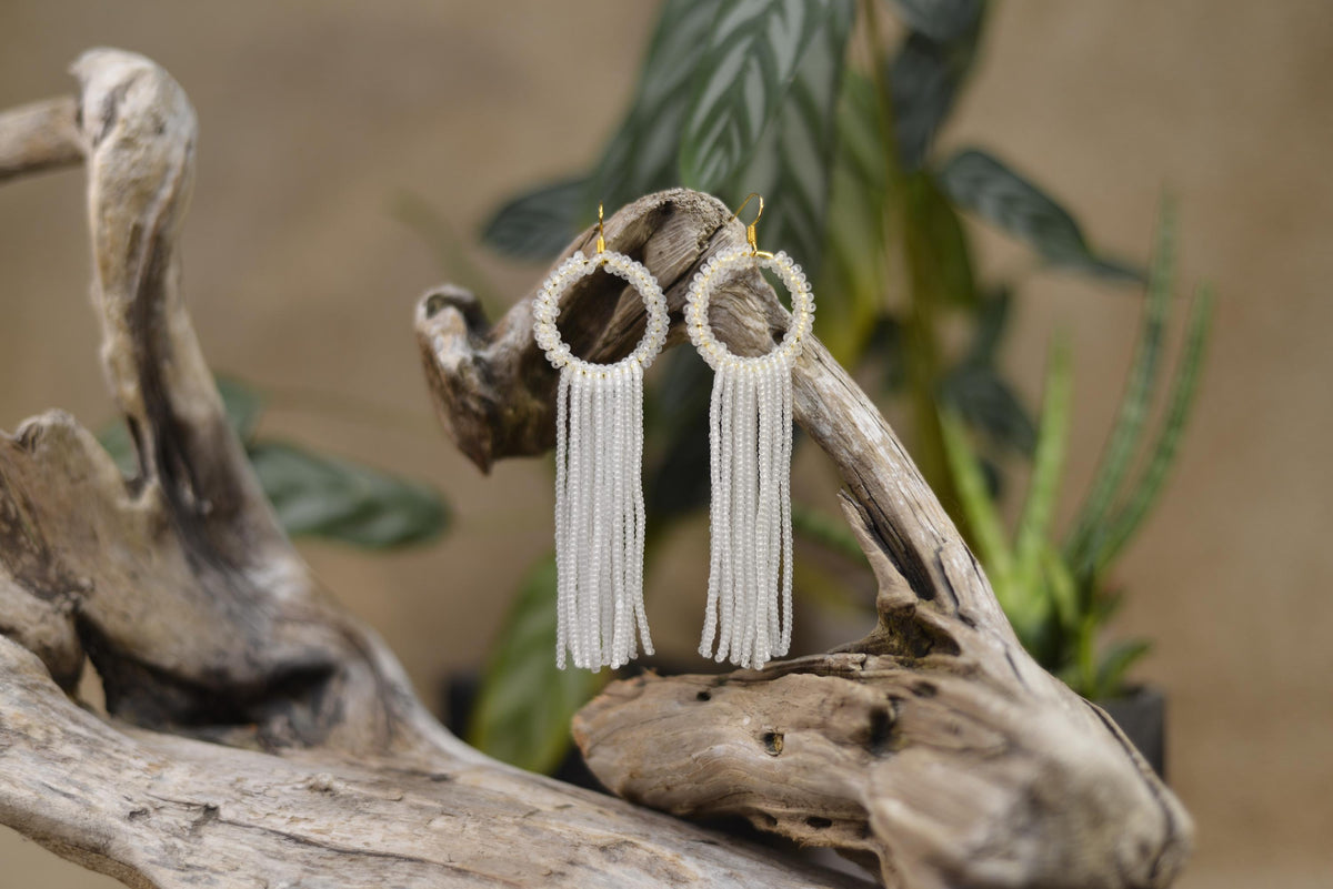 A pair of white earrings with tassels hanging from a wooden branch, placed on a plant stem.