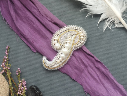 a decorative piece of jewelry, possibly a brooch or pin, with a purple fabric draped over it. The jewelry is adorned with pearls and rhinestones, giving it an elegant and sophisticated appearance. The background includes a gray fabric and some feathers, adding a touch of nature and texture to the overall composition.
