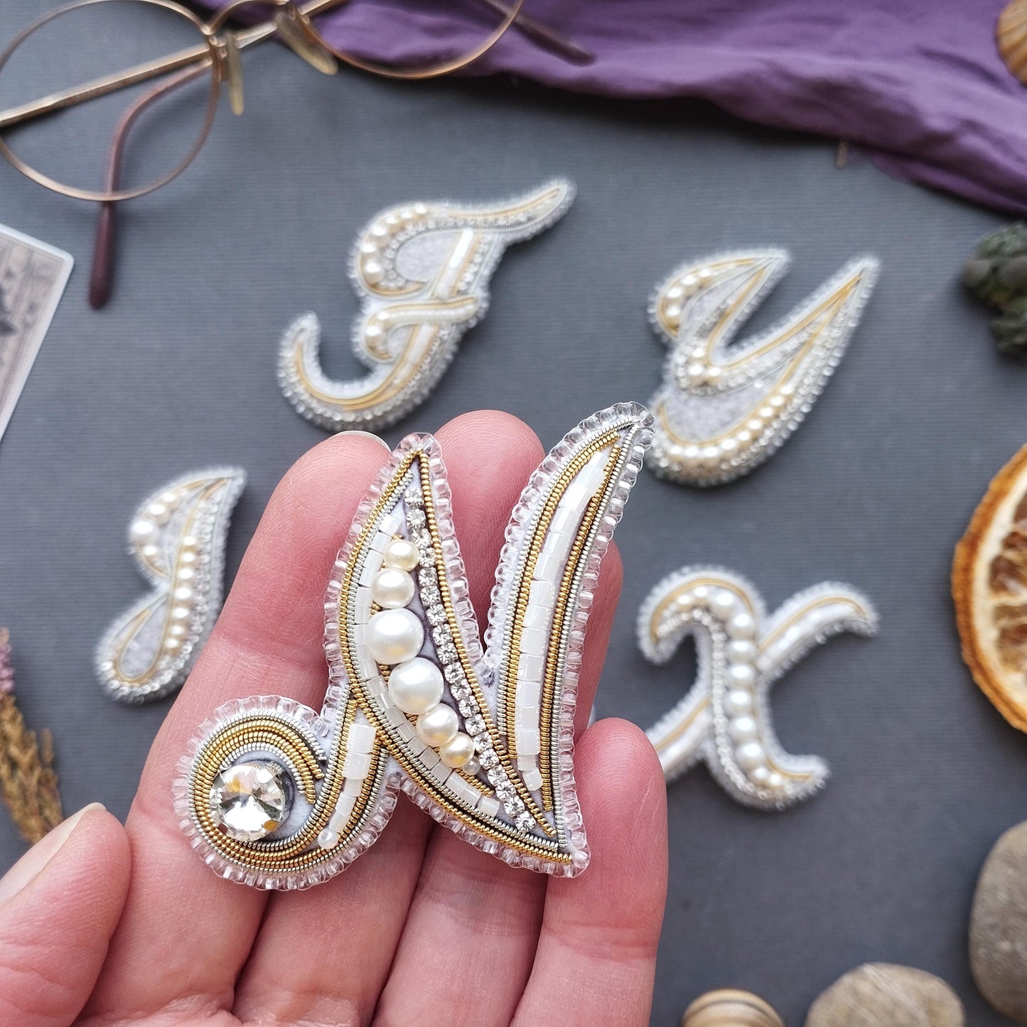 a hand holding a decorative letter n made of beads and pearls, with other decorative elements such as pearls and a pair of glasses in the background.