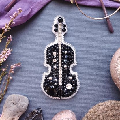 a black and white beaded violin brooch, surrounded by various objects such as a pair of glasses, a flower, and a rock.