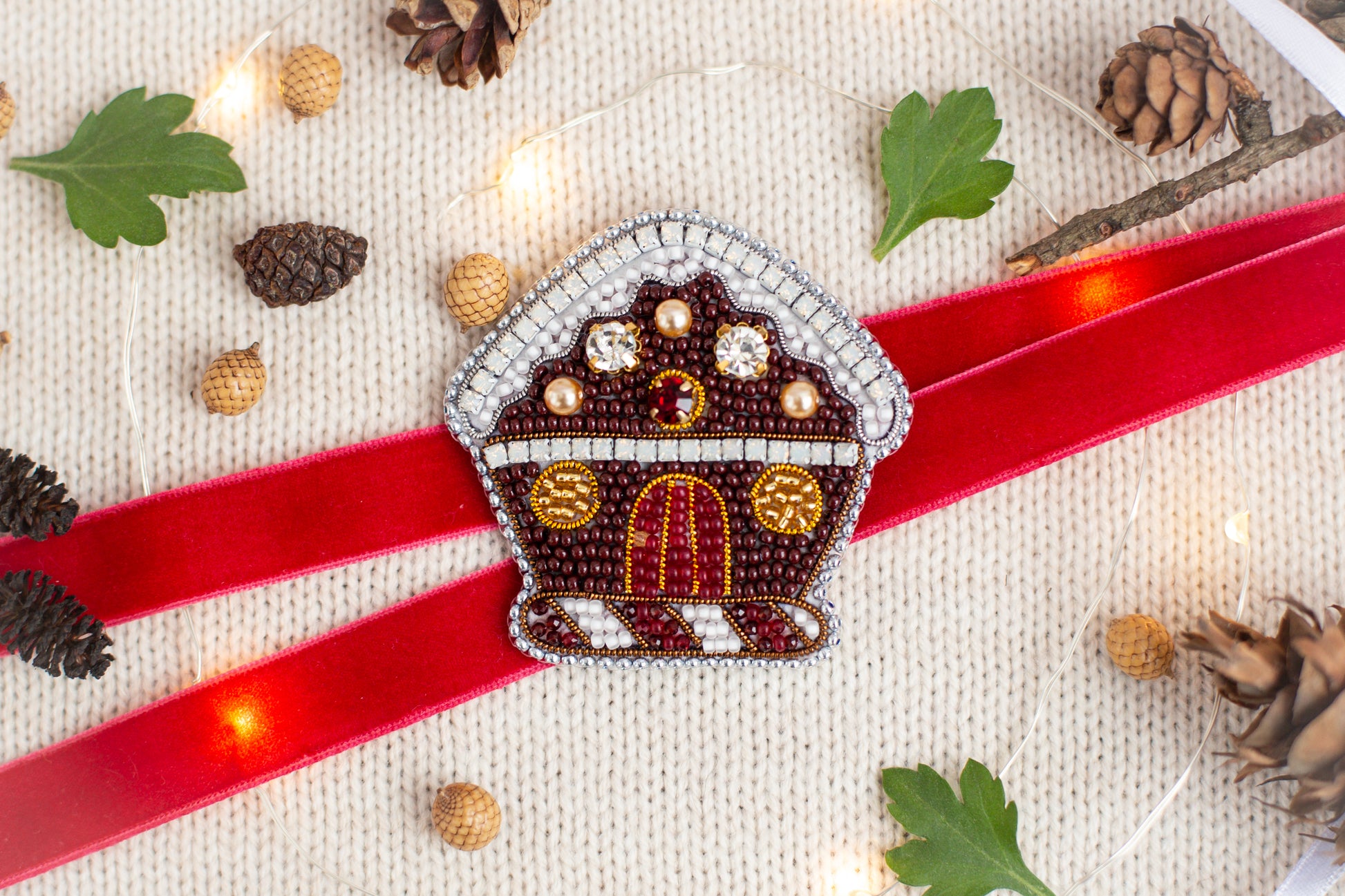 a red ribbon with a beaded house-shaped ornament on it, surrounded by pine cones, leaves, and Christmas lights.