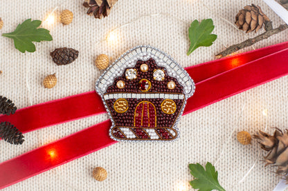 a red ribbon with a beaded house-shaped ornament on it, surrounded by pine cones, leaves, and Christmas lights.