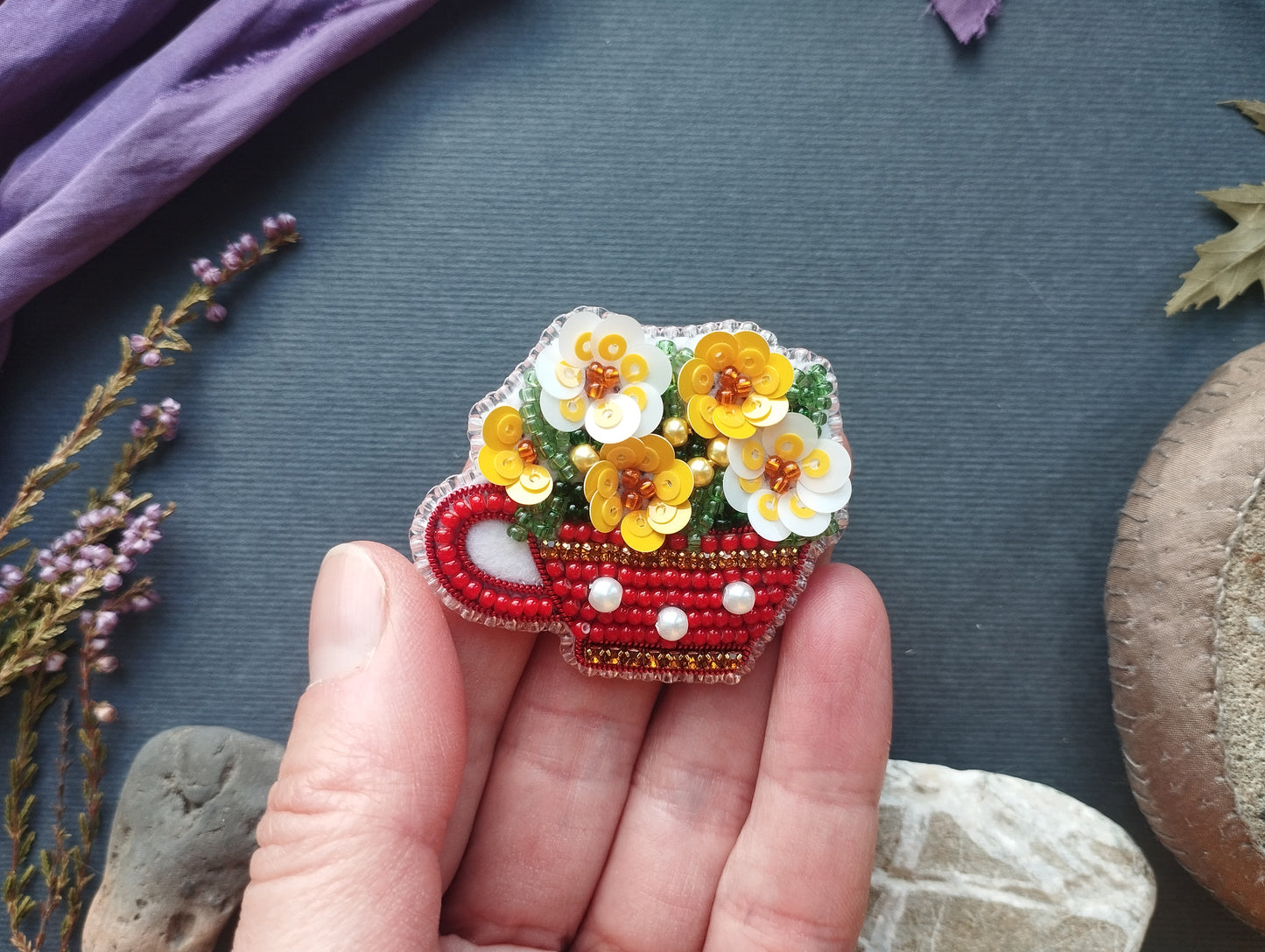 A hand holding a small, intricately beaded brooch in the shape of a cup, with yellow flowers on top.
