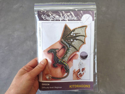 A hand holding a package containing a kit for creating a green dragon embroidery piece.