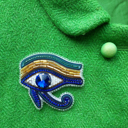 a close-up view of a green fabric with a decorative eye-shaped brooch adorned with blue and gold beads, a large blue gemstone, and a small green ball.