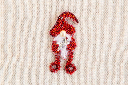 A red ornament resembling a gnome or elf, made of beads and wire, is displayed against a beige background.