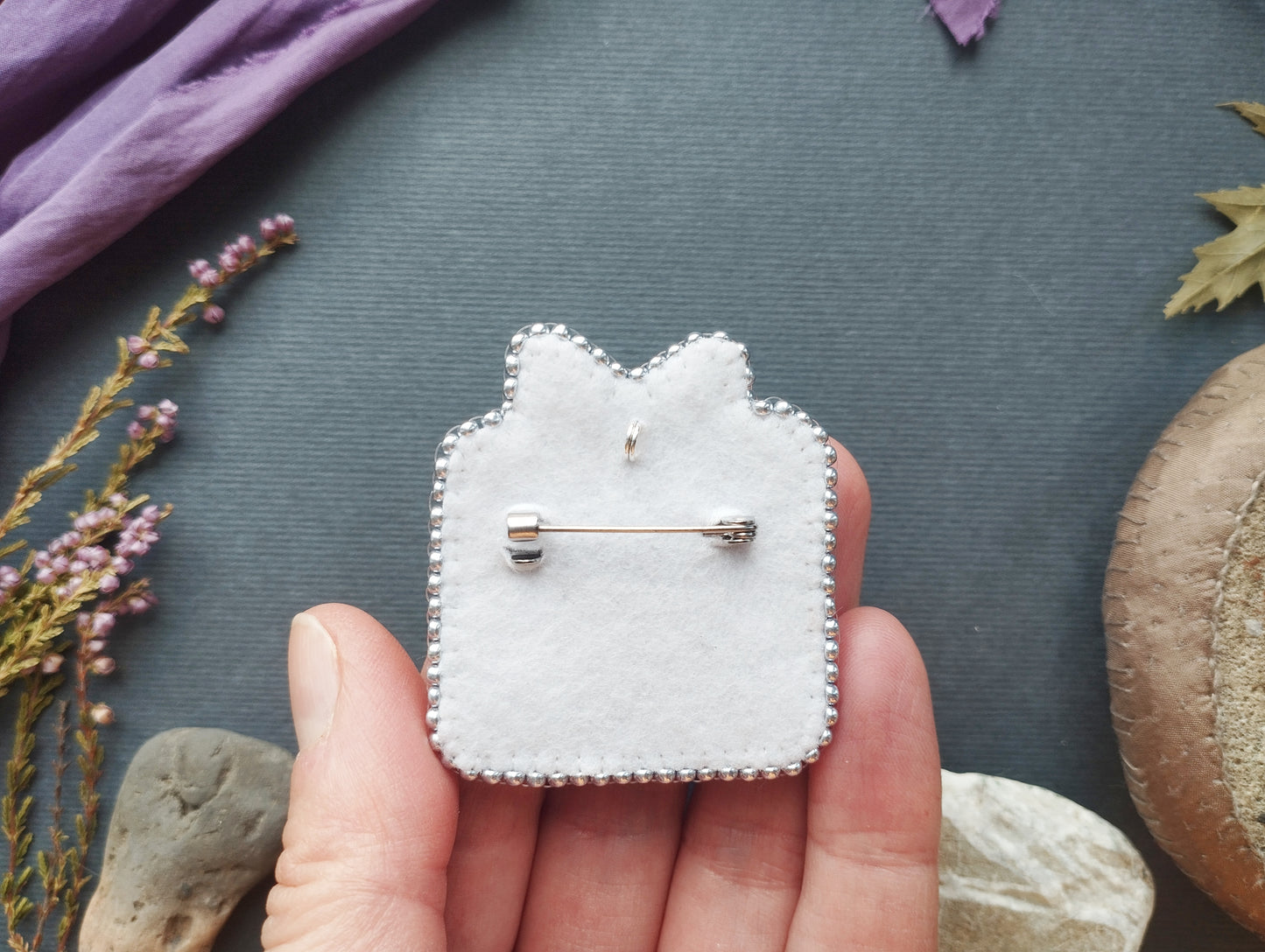 A hand holding a white felt pin with a silver pinback design, surrounded by various natural elements such as flowers, rocks, and leaves.