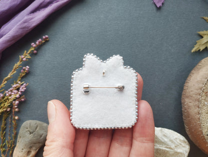A hand holding a white felt pin with a silver pinback design, surrounded by various natural elements such as flowers, rocks, and leaves.