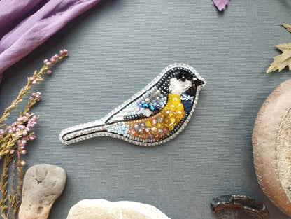 a decorative bird embroidered with colorful beads, surrounded by various natural elements such as leaves, flowers, and rocks.