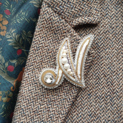 A close-up image of a decorative brooch or pin with a floral pattern and a crystal or gemstone at the center, attached to a tweed or woolen fabric.