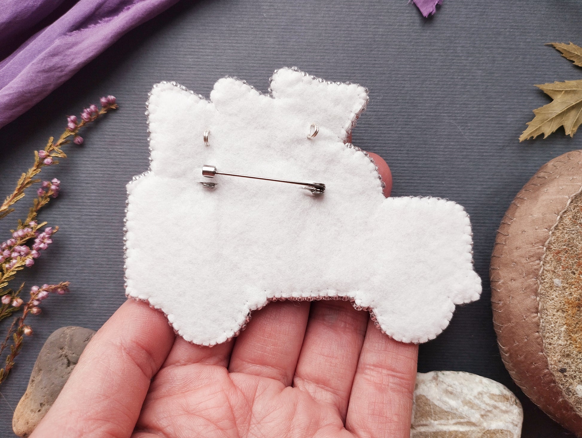 A hand holding a white felt object resembling a pin or badge, with a small pin or button attached to it. The hand is positioned over a variety of objects, including a purple fabric, leaves, and rocks, creating a natural and outdoor-themed setting.