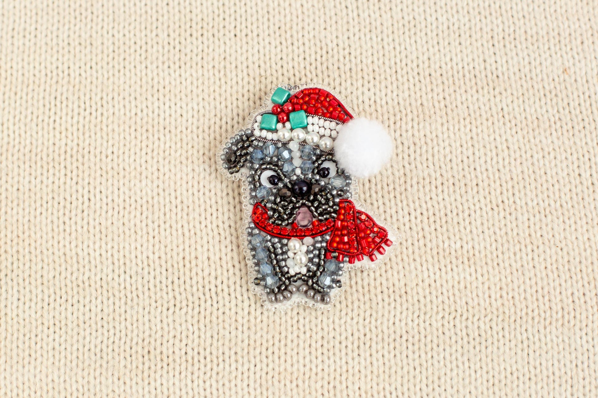 A small, intricately beaded dog wearing a red Santa hat and scarf is displayed on a beige textured background.