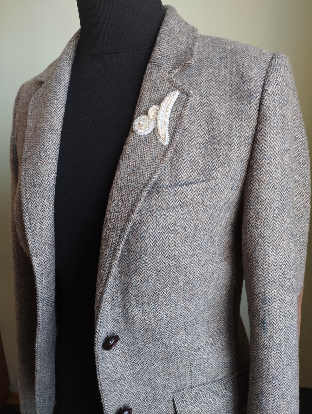 A gray tweed blazer with a white decorative pin on the lapel, displayed on a mannequin.