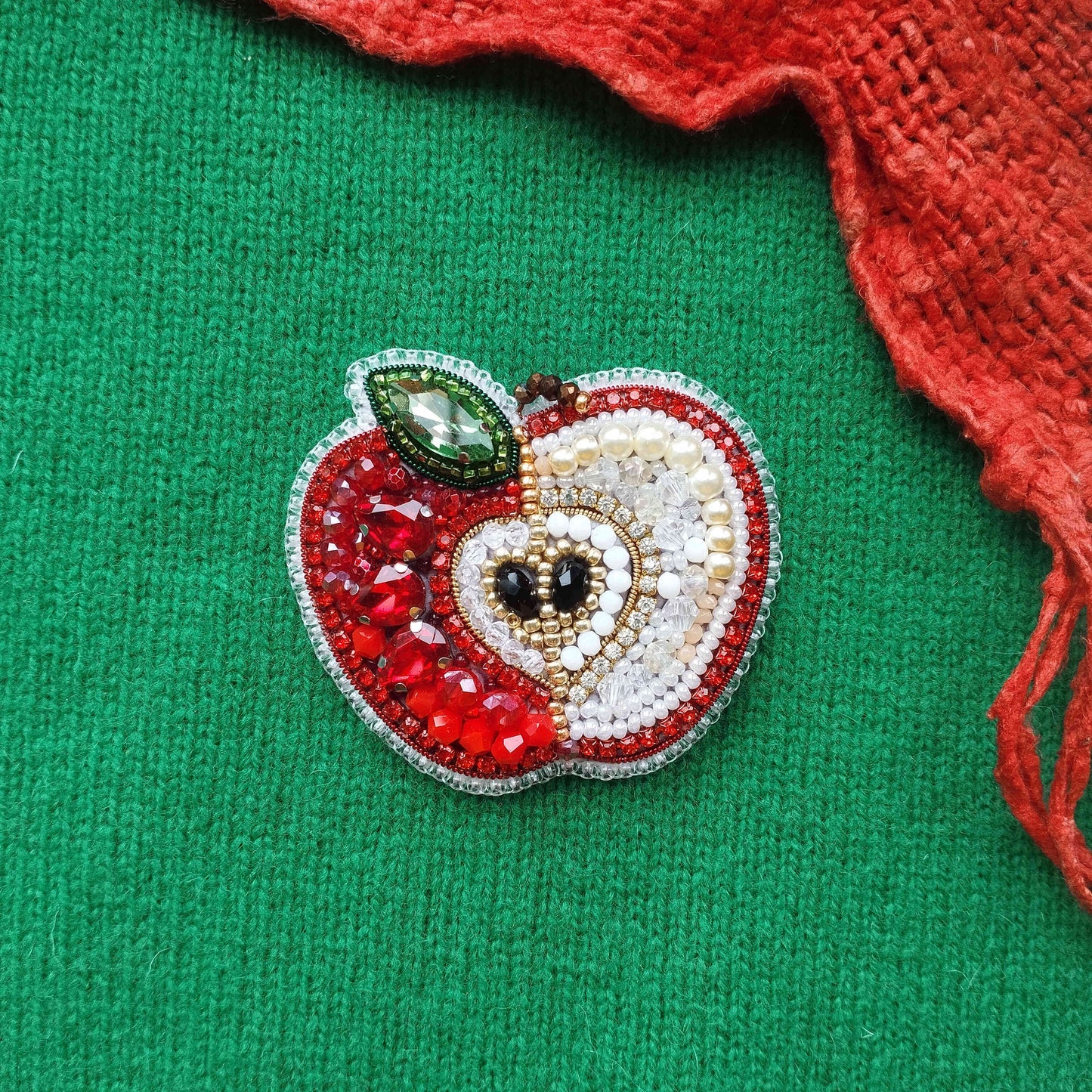A decorative apple-shaped brooch made of beads, gemstones, and a leaf, placed on a green fabric background.