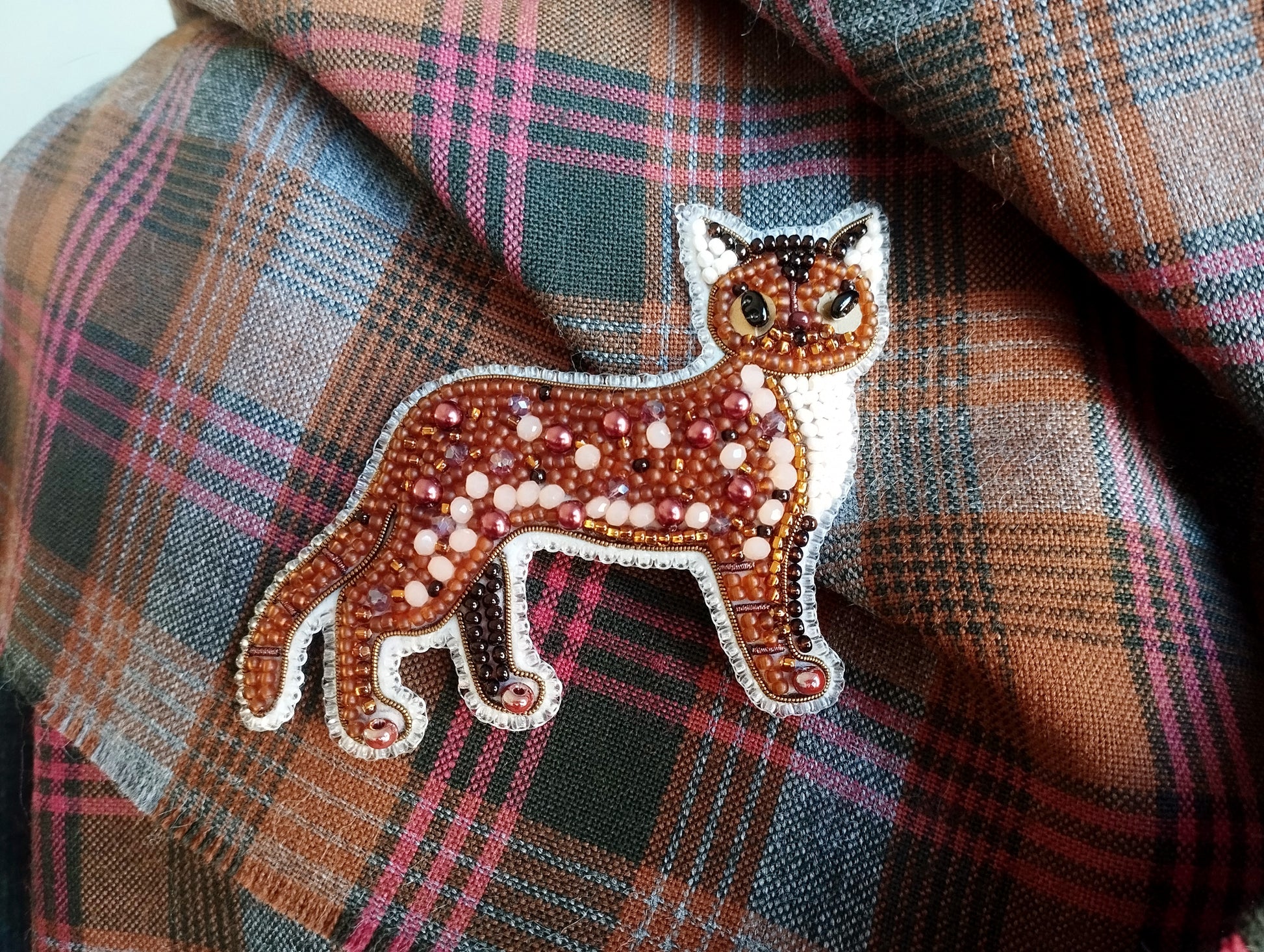 A close-up image of a decorative beaded cat pin or brooch placed on a plaid fabric background.