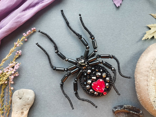 a detailed close-up of a black spider-shaped brooch adorned with red gemstones and black beads, resting on a gray surface.