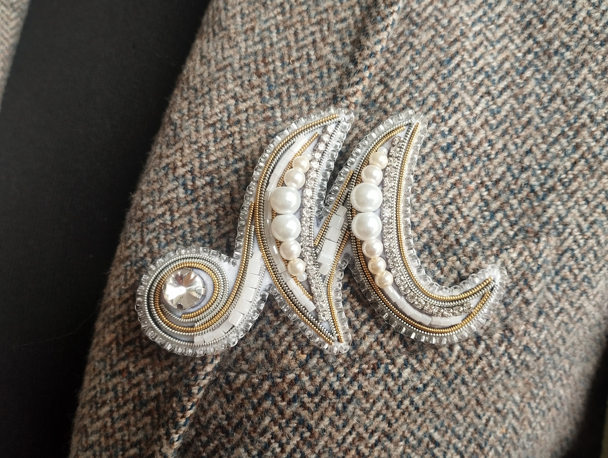 A close-up image of a decorative monogram brooch or pin, featuring a combination of pearls, diamonds, and a crystal element.