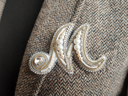 A close-up image of a decorative monogram brooch or pin, featuring a combination of pearls, diamonds, and a crystal element.