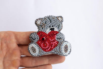 a hand holding a beaded teddy bear with a heart