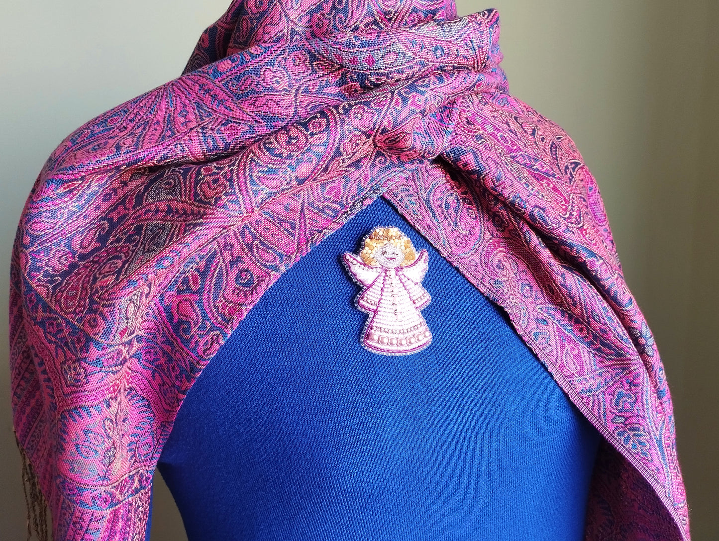 A close-up of a person wearing a blue shirt with a pink and purple paisley patterned scarf draped over their shoulders. A small, intricately designed pin or brooch is visible on the person's chest.