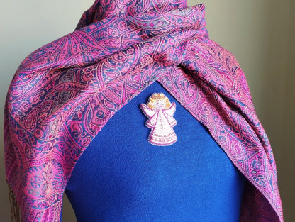 A close-up of a person wearing a blue shirt with a pink and purple paisley patterned scarf draped over their shoulders. A small, intricately designed pin or brooch is visible on the person's chest.