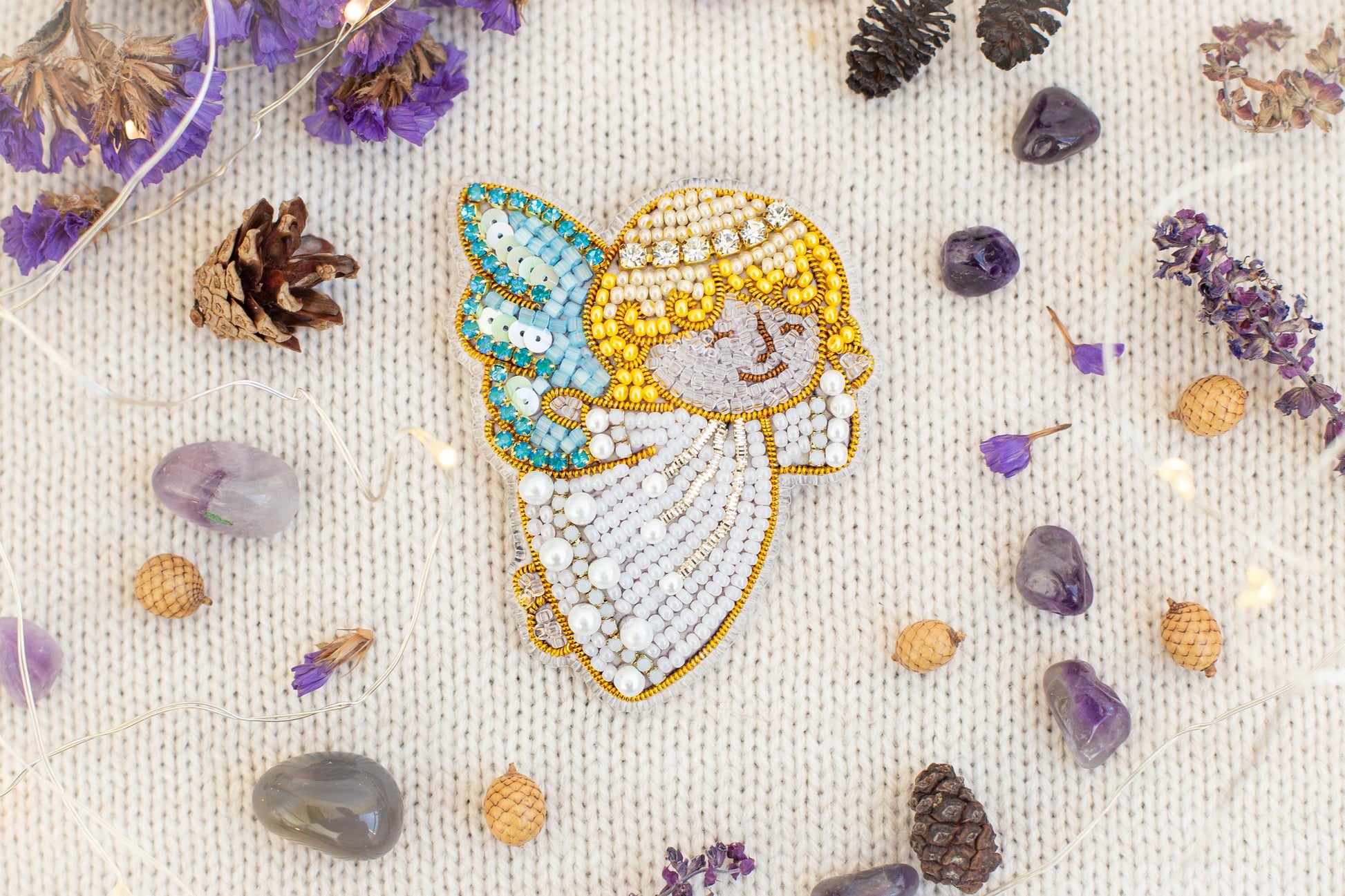 a close-up of a beaded embroidered figure surrounded by various natural elements, including pine cones, gemstones, and purple flowers.