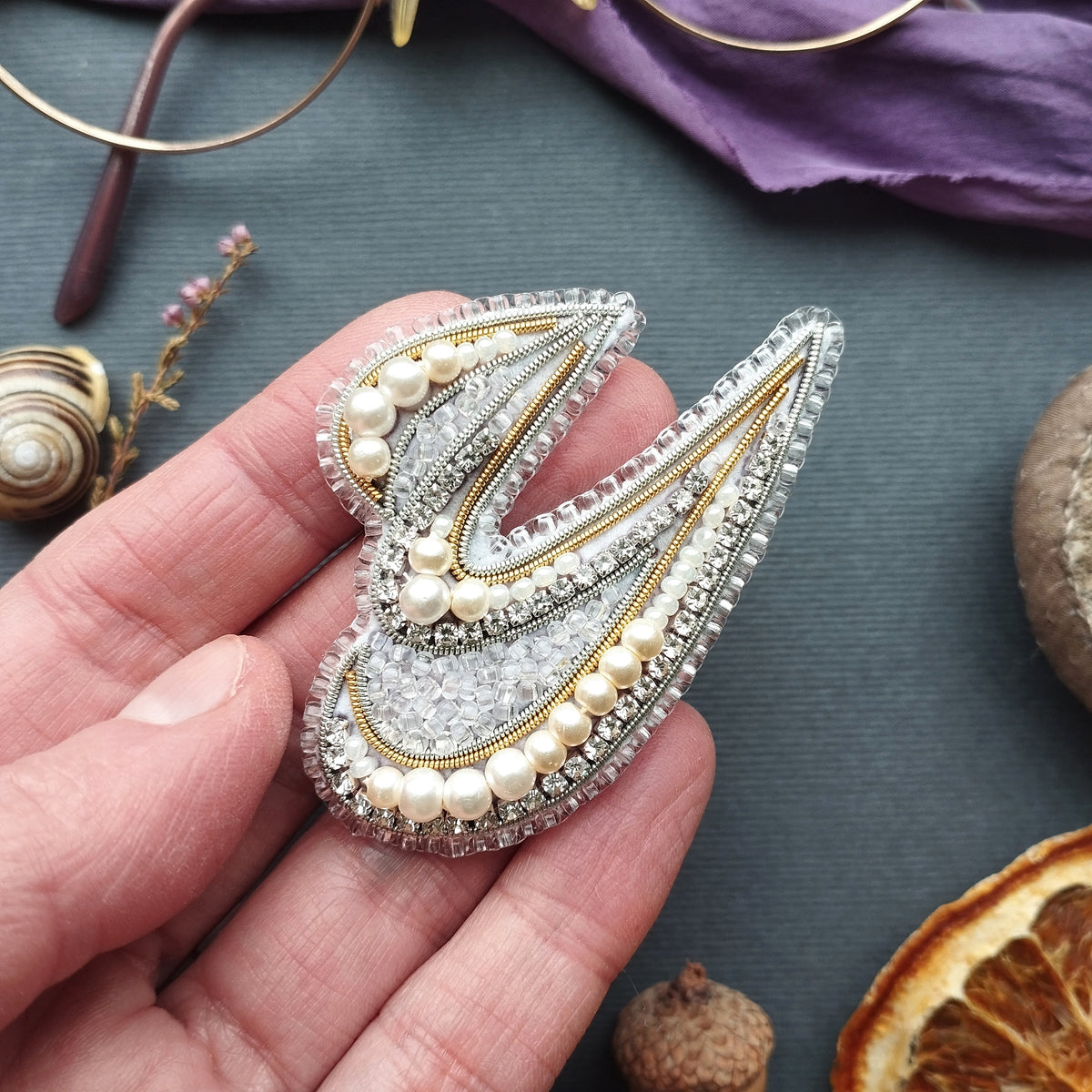 A hand holding a decorative beaded brooch with a floral design, surrounded by various natural elements such as a seashell, dried flowers, and an orange slice.