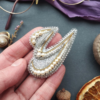 A hand holding a decorative beaded brooch with a floral design, surrounded by various natural elements such as a seashell, dried flowers, and an orange slice.