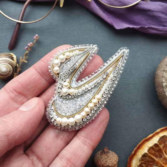 A hand holding a decorative beaded brooch with a floral design, surrounded by various natural elements such as a seashell, dried flowers, and an orange slice.
