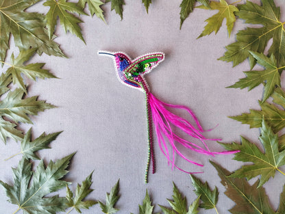 a colorful hummingbird brooch surrounded by green leaves on a gray background.