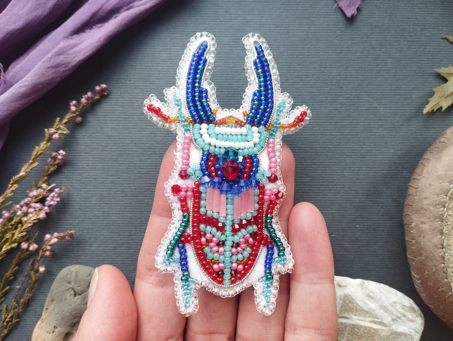 A hand holding a colorful, intricately beaded insect-shaped object against a dark background.