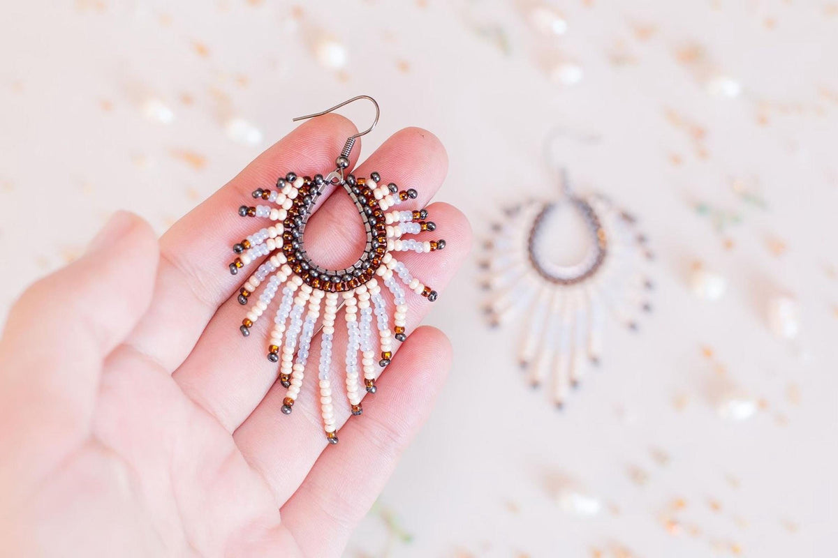 A hand holding a pair of earrings with a beaded design, against a blurred background of more earrings.