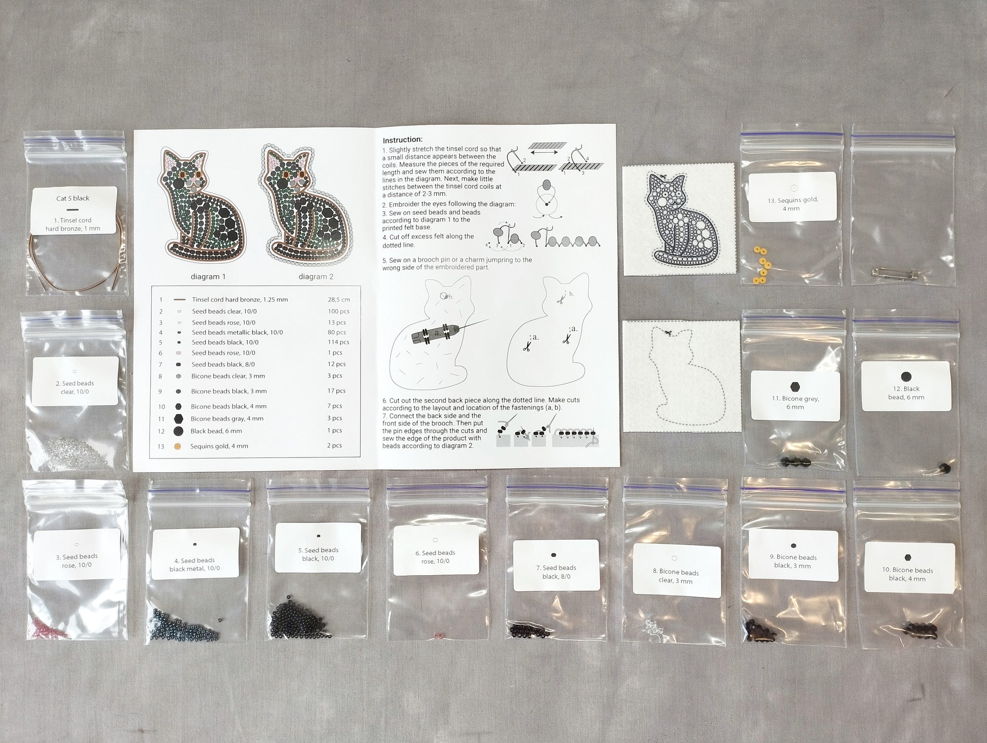 various craft supplies, including bags of beads, seeds, and other materials, arranged on a table. There is also a printed guide or instructions sheet for a project involving cat-themed items, such as earrings and cat-shaped objects.