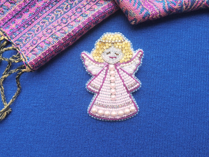 A hand-beaded angel figure in a pink dress is displayed on a blue background, with a colorful fabric or textile visible in the background.